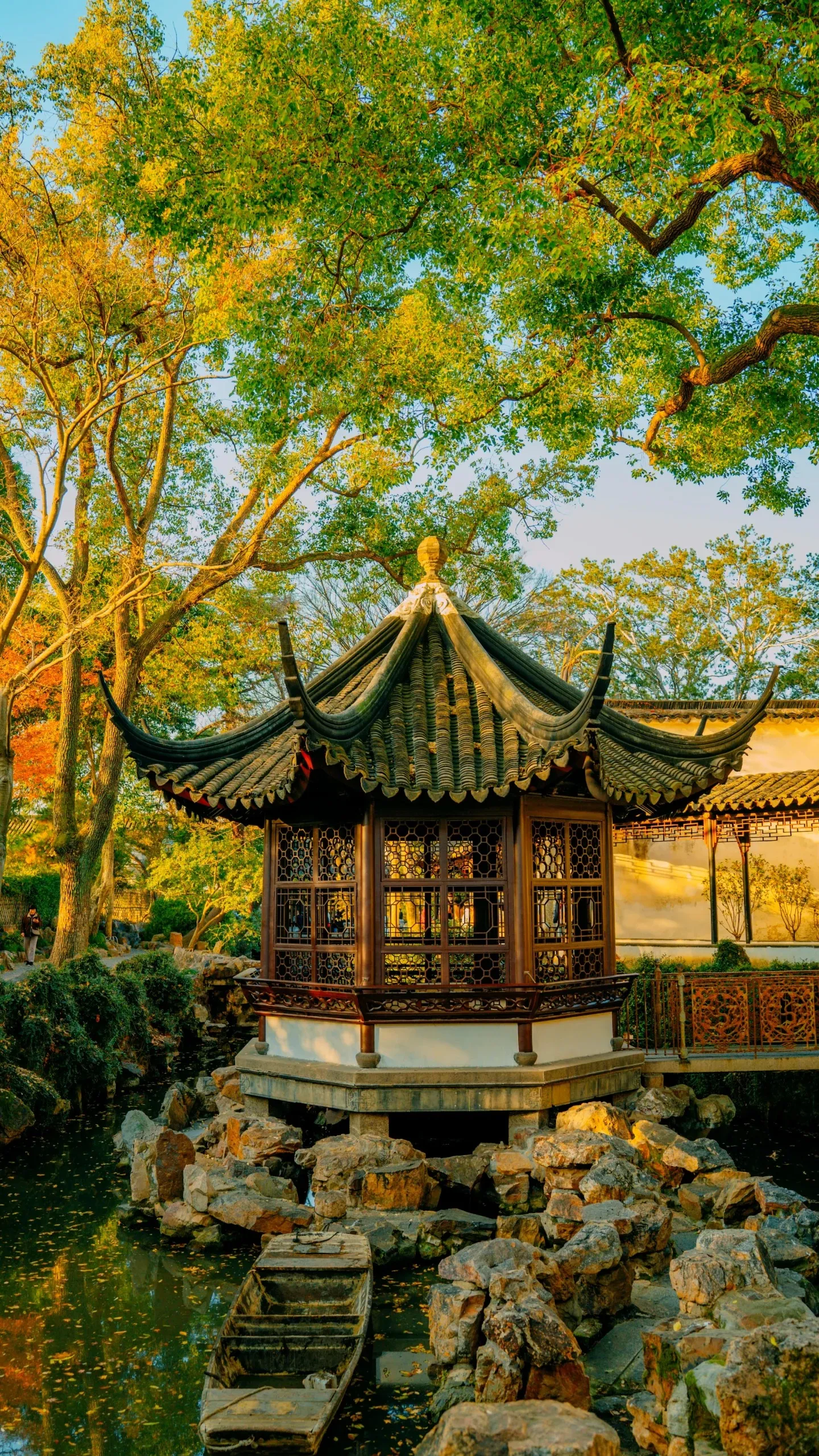 A pavilion with a green tiled roof standing on a rockery above a pond in the Humble Administrator's Garden, Suzhou, a wooden boat moored below.
